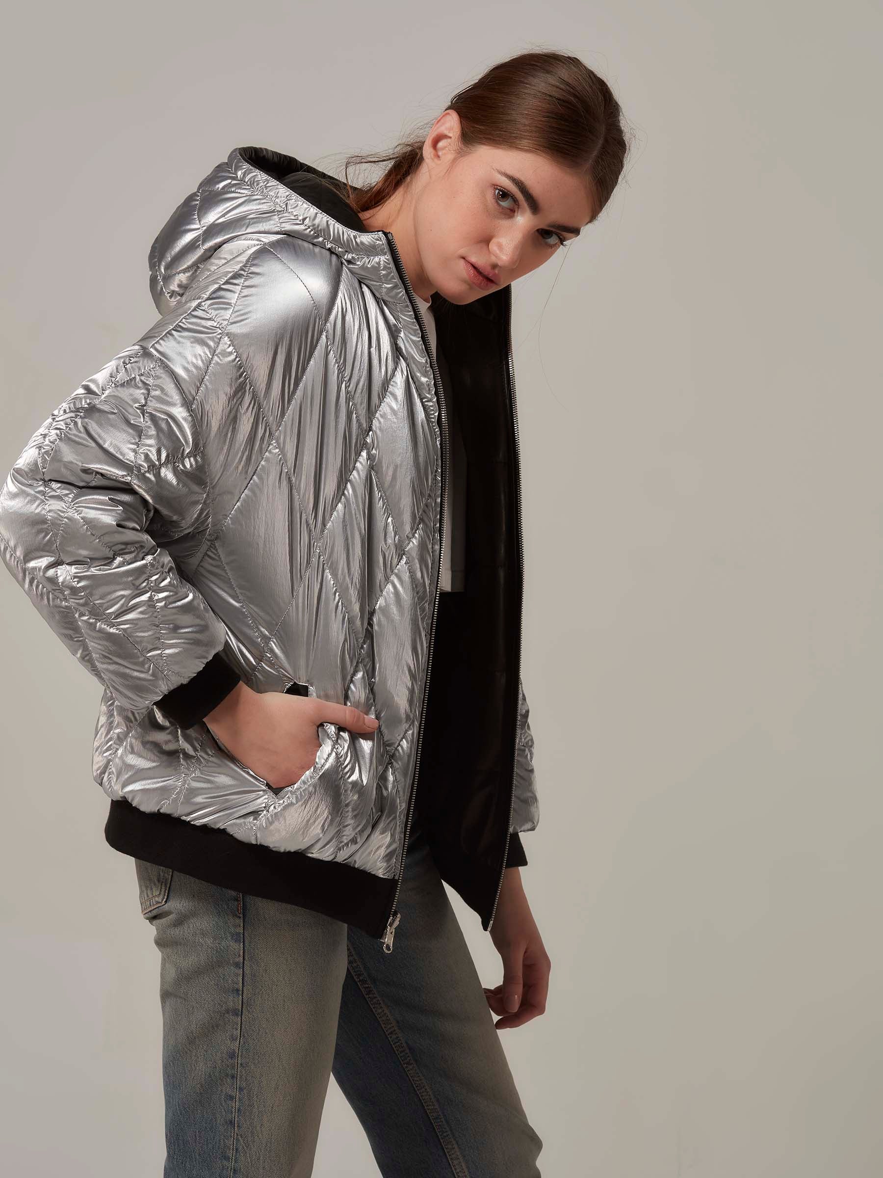 Close-up front view of a stylish woman in a black lambskin leather and metallic silver waterproof fabric double-sided reversible puffer jacket. She is wearing fabric side and poses with the front zipper open, exuding a rebellious vibe.