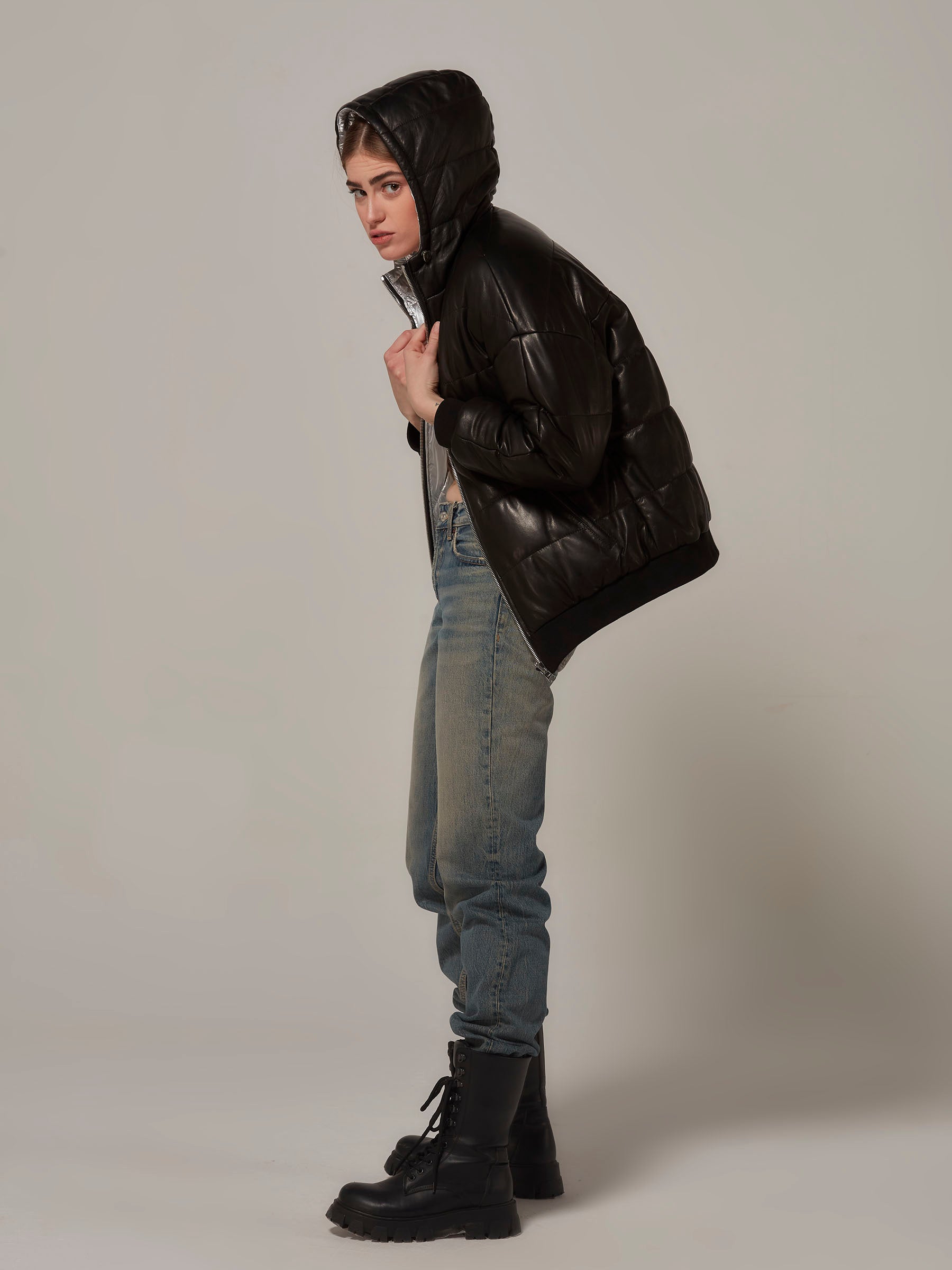 A full-length photo of a stylish woman in a black leather and metallic silver reversible puffer jacket. She poses with a rebellious vibe, holding the collar from both sides. Side view.