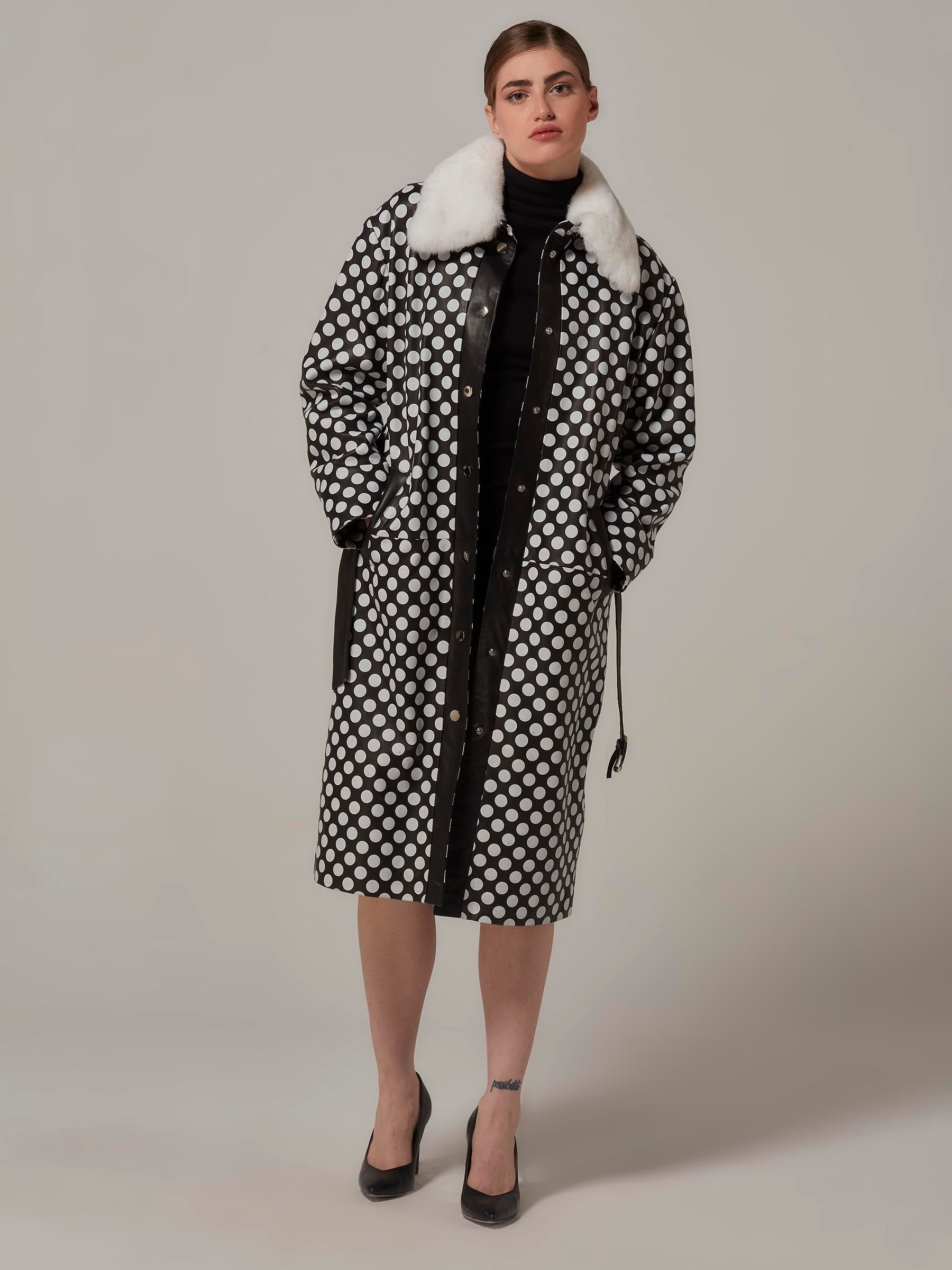 Front view of a coolly posed woman wearing a Black and White Leather Coat with Polka Dot and Rex Fur Collar, hands in pockets. Full legth photo.